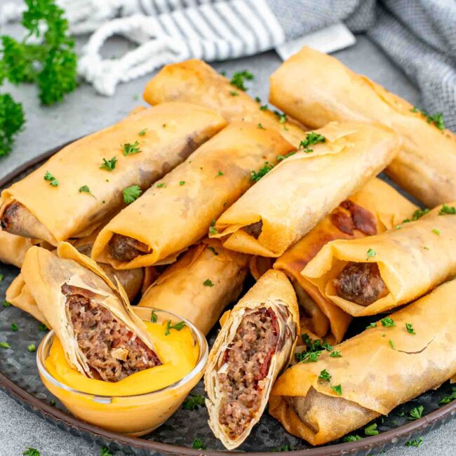 bacon cheeseburger egg rolls on a metal plate along a small bowl with cheese sauce.