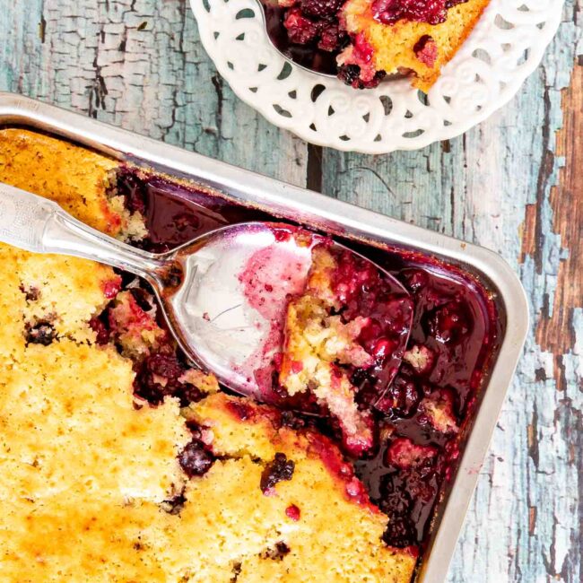 freshly baked blackberry cobbler in a baking pan with a couple servings missing and a serving spoon inside.