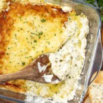 Creamy, cheesy roasted garlic dip with a golden top, partially scooped with a wooden spoon, served alongside crusty bread.