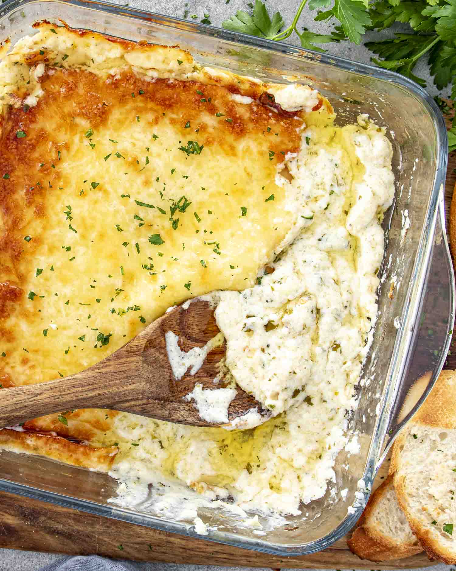 Creamy, cheesy roasted garlic dip with a golden top, partially scooped with a wooden spoon, served alongside crusty bread.