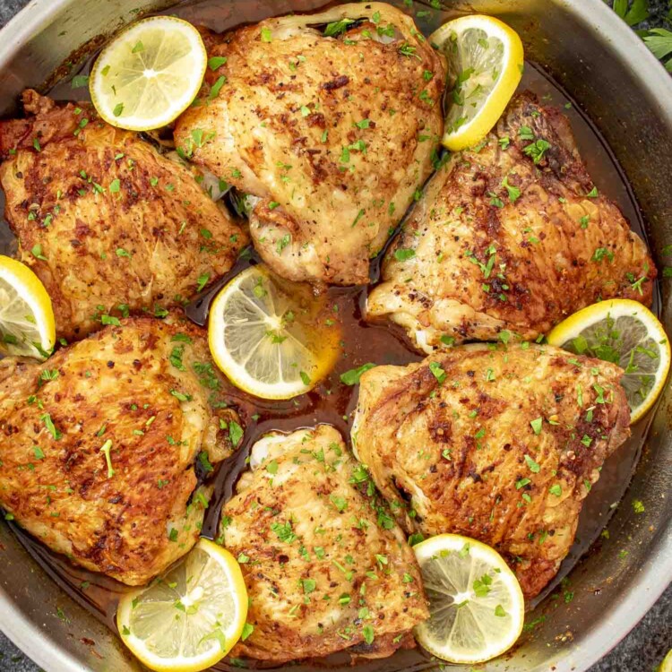 crispy lemon chicken thighs fresh out of the oven in a skillet garnished with parsley and lemon slices.