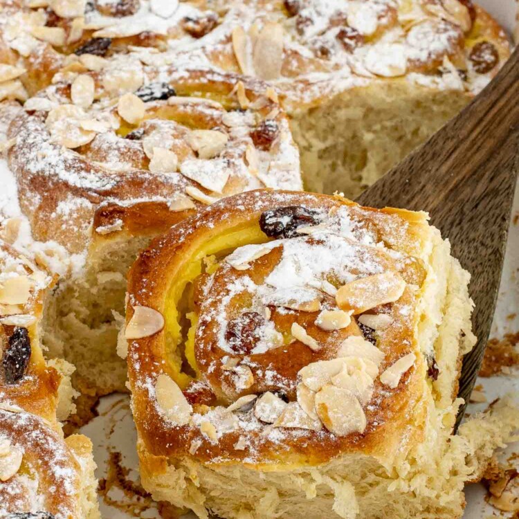 a spatula lifting a custard raisin roll from a pan full of rolls, dusted with a bit of icing sugar.