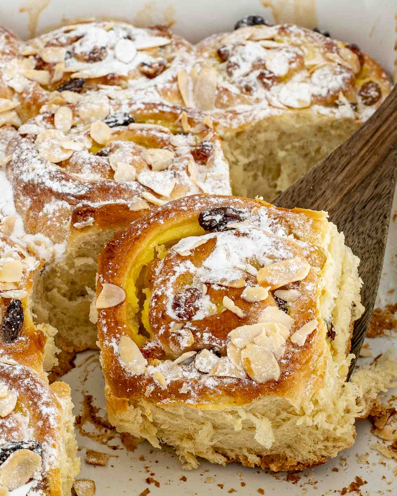 a spatula lifting a custard raisin roll from a pan full of rolls, dusted with a bit of icing sugar.