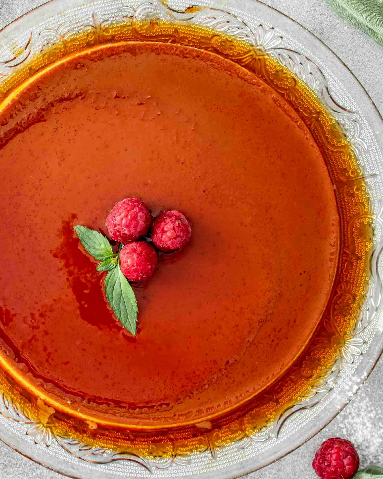a silky smooth flan, glossy caramel top, garnished with fresh raspberries on a glass platter.