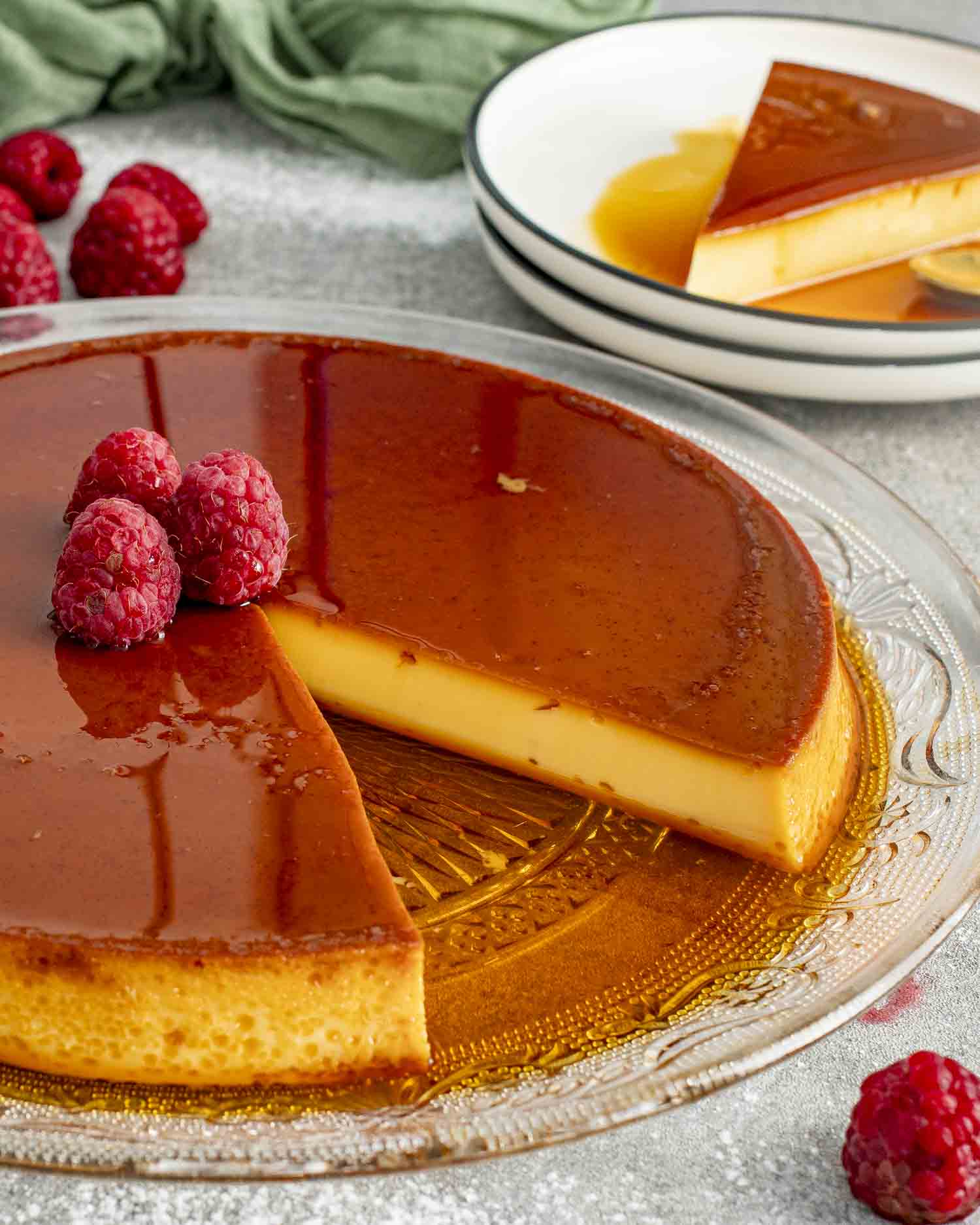 A beautifully presented flan with a smooth, glossy caramel top, garnished with fresh raspberries on a glass platter. One slice is removed, showing the creamy, rich custard interior.