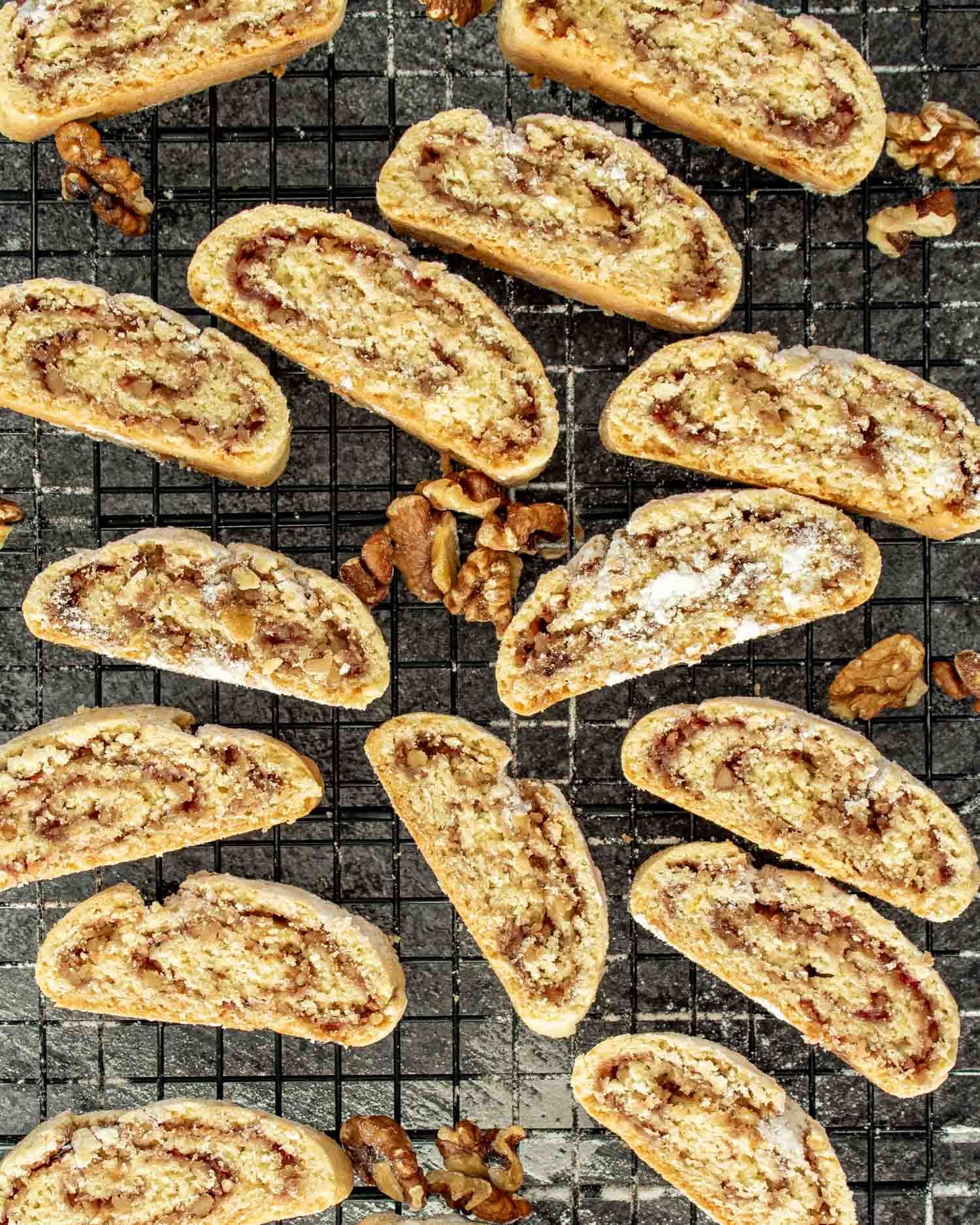 Freshly baked Italian nut roll cookies cooling on a wire rack, dusted with powdered sugar and surrounded by scattered walnut pieces.