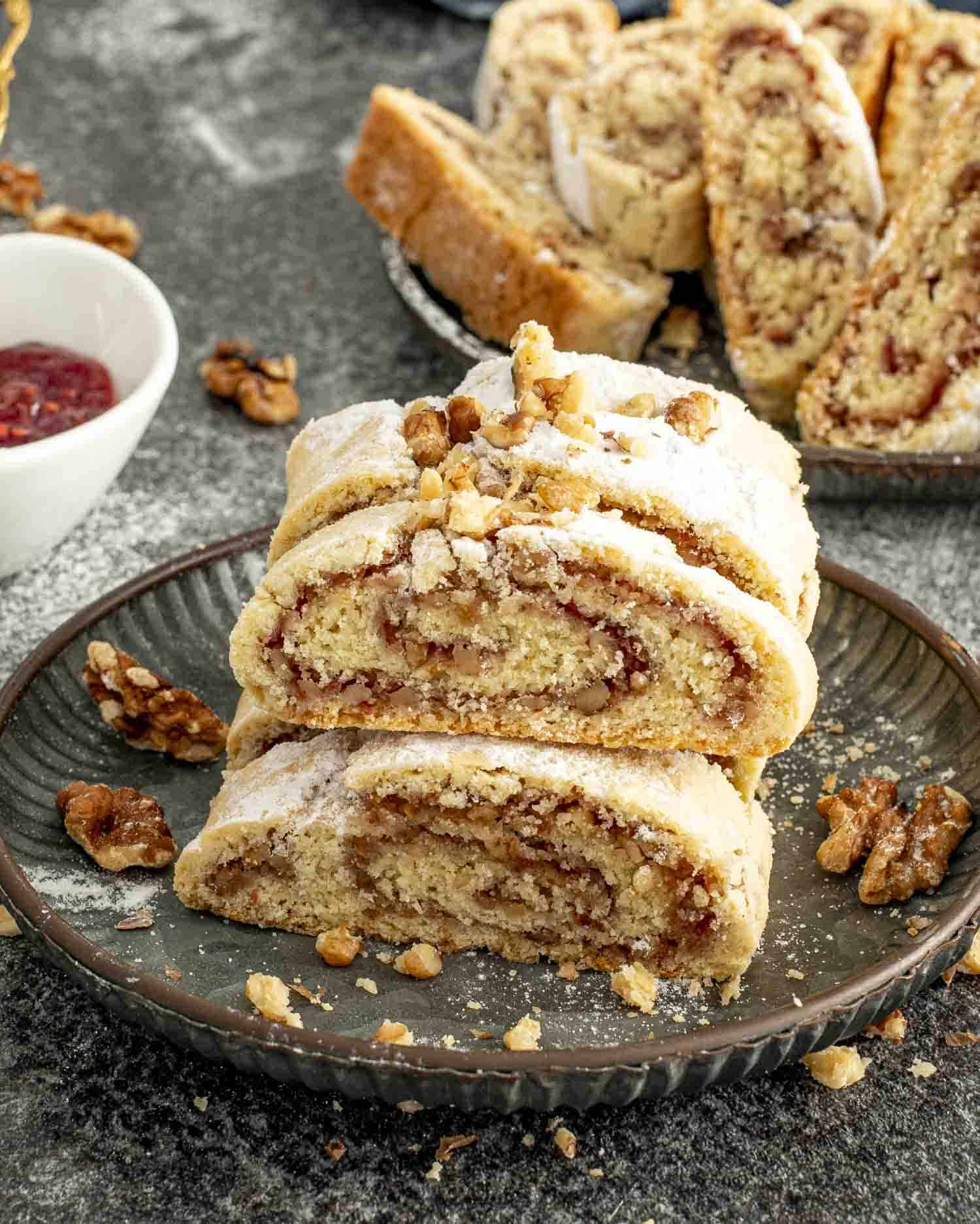 Close-up of sliced Italian nut roll cookies on a dark plate, showcasing a nutty jam filling, flaky texture, and dusting of powdered sugar.