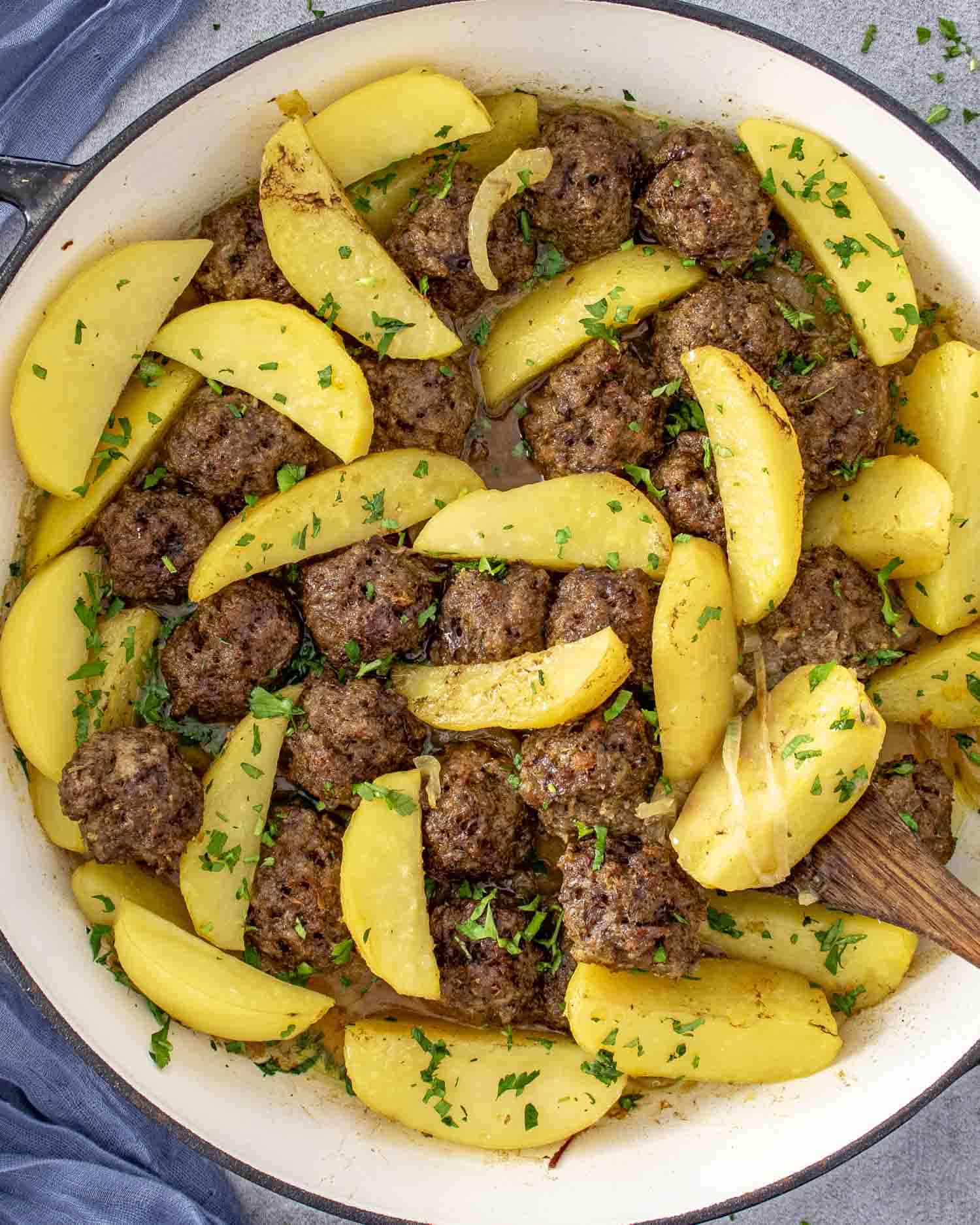 A close-up of a large white pot filled with golden potato wedges and browned meatballs, garnished with fresh chopped parsley. The dish is saucy, vibrant, and comforting, showcasing a hearty one-pot meal.