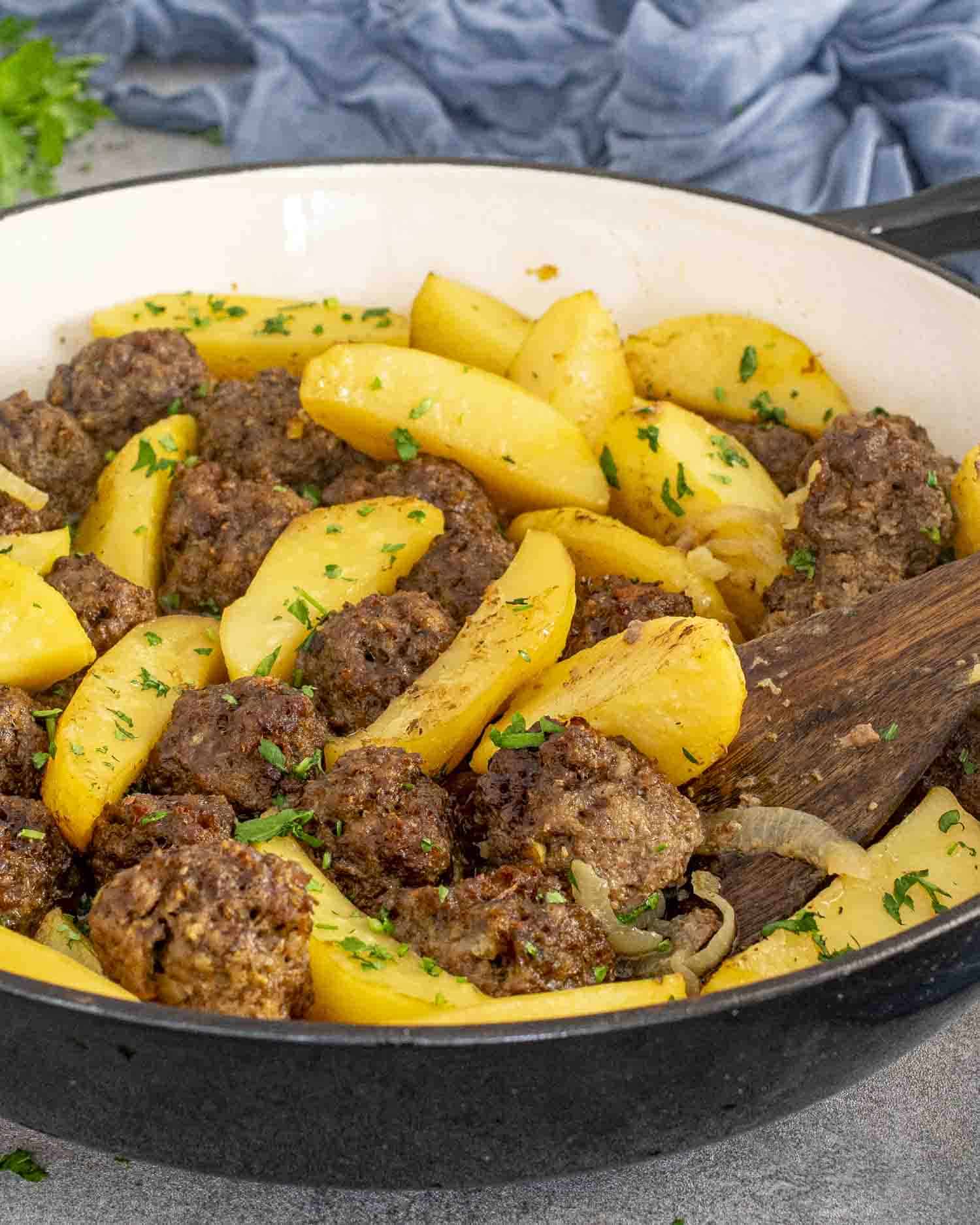 A close-up of a large white pot filled with golden potato wedges and browned meatballs, garnished with fresh chopped parsley. The dish is saucy, vibrant, and comforting, showcasing a hearty one-pot meal.