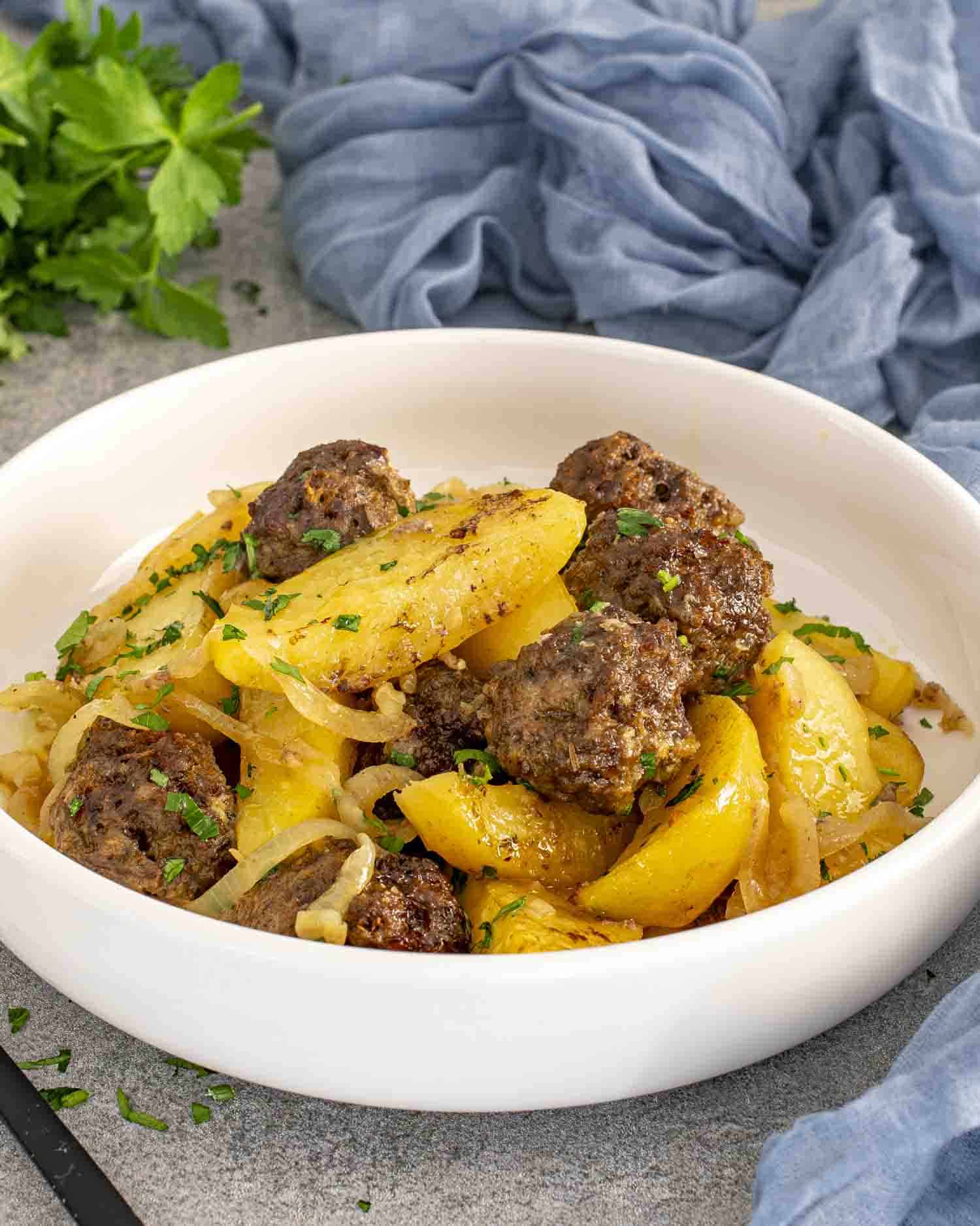 A white bowl filled with a serving of golden potato wedges and juicy meatballs, garnished with fresh parsley. The dish is hearty and vibrant, with caramelized onions adding a touch of warmth, sitting on a textured gray surface with a blue cloth in the background.