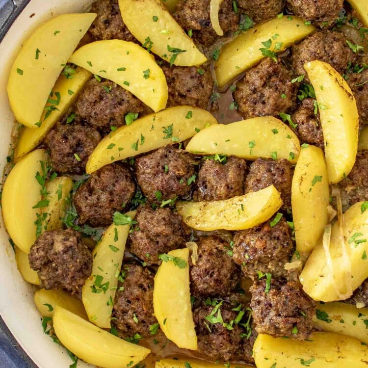 A close-up of a large white pot filled with golden potato wedges and browned meatballs, garnished with fresh chopped parsley. The dish is saucy, vibrant, and comforting, showcasing a hearty one-pot meal.