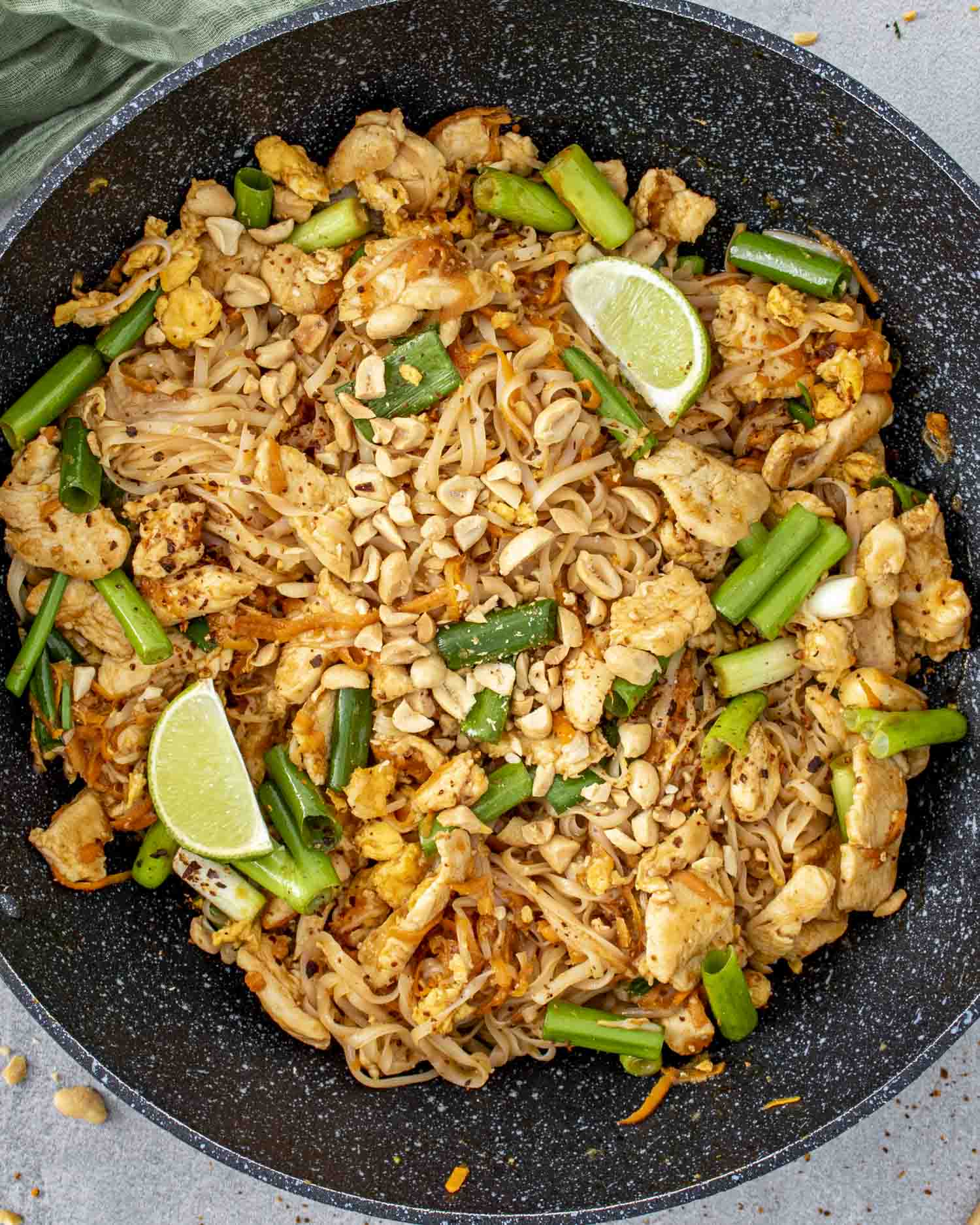 A skillet filled with freshly made Pad Thai featuring rice noodles, tender chicken, green onions, scrambled eggs, and crushed peanuts. Lime wedges are nestled on top for garnish, with vibrant green onions scattered throughout the dish. 