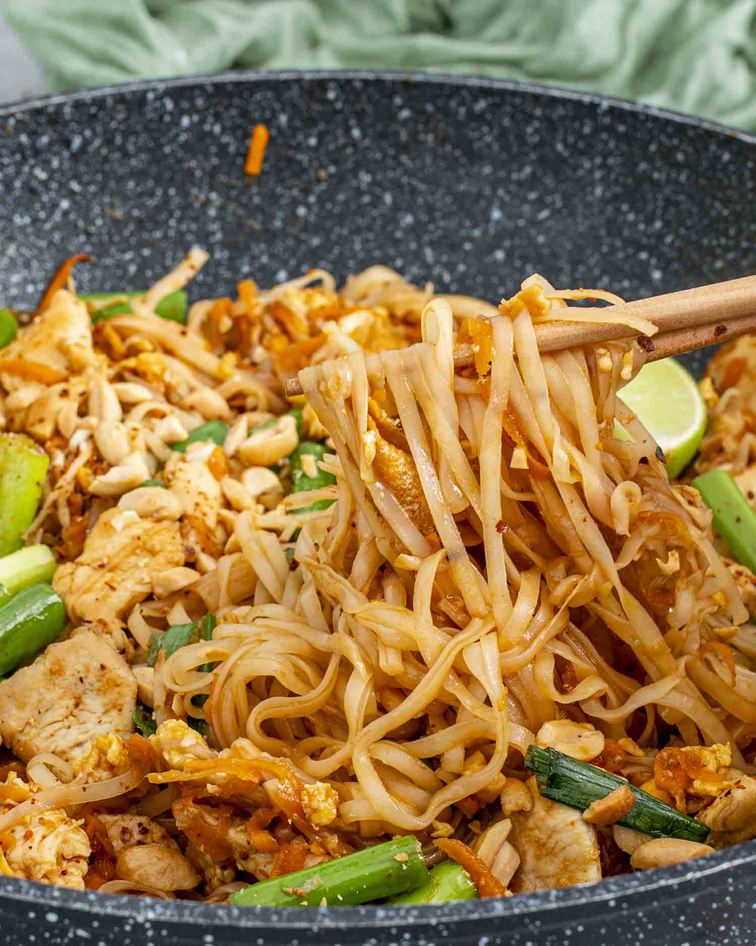 A skillet filled with freshly made Pad Thai featuring rice noodles, tender chicken, green onions, scrambled eggs, and crushed peanuts. Lime wedges are nestled on top for garnish, with vibrant green onions scattered throughout the dish.