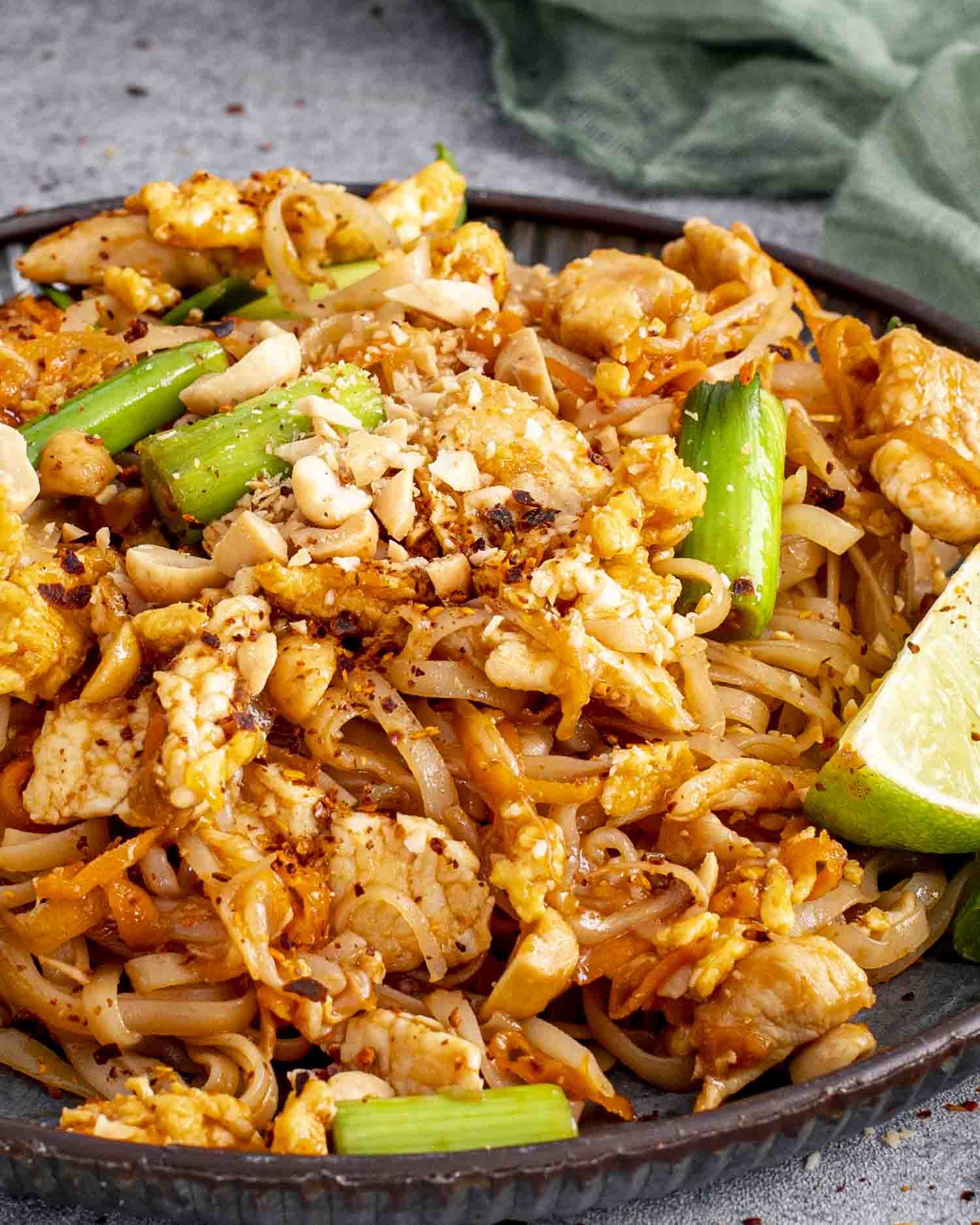 A plate of Pad Thai with rice noodles, tender chicken, scrambled eggs, green onions, and crushed peanuts. Chopsticks are lifting a portion of noodles, while a lime wedge adds a pop of color on the side.