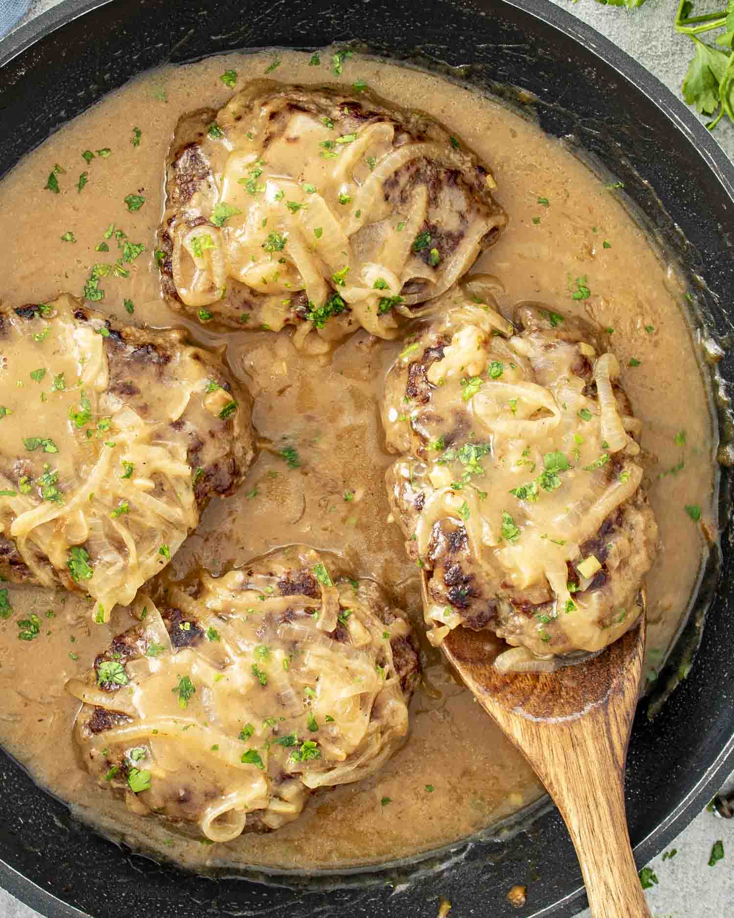 Juicy beef patties covered in rich, creamy onion gravy, garnished with fresh parsley, served hot in a black skillet.