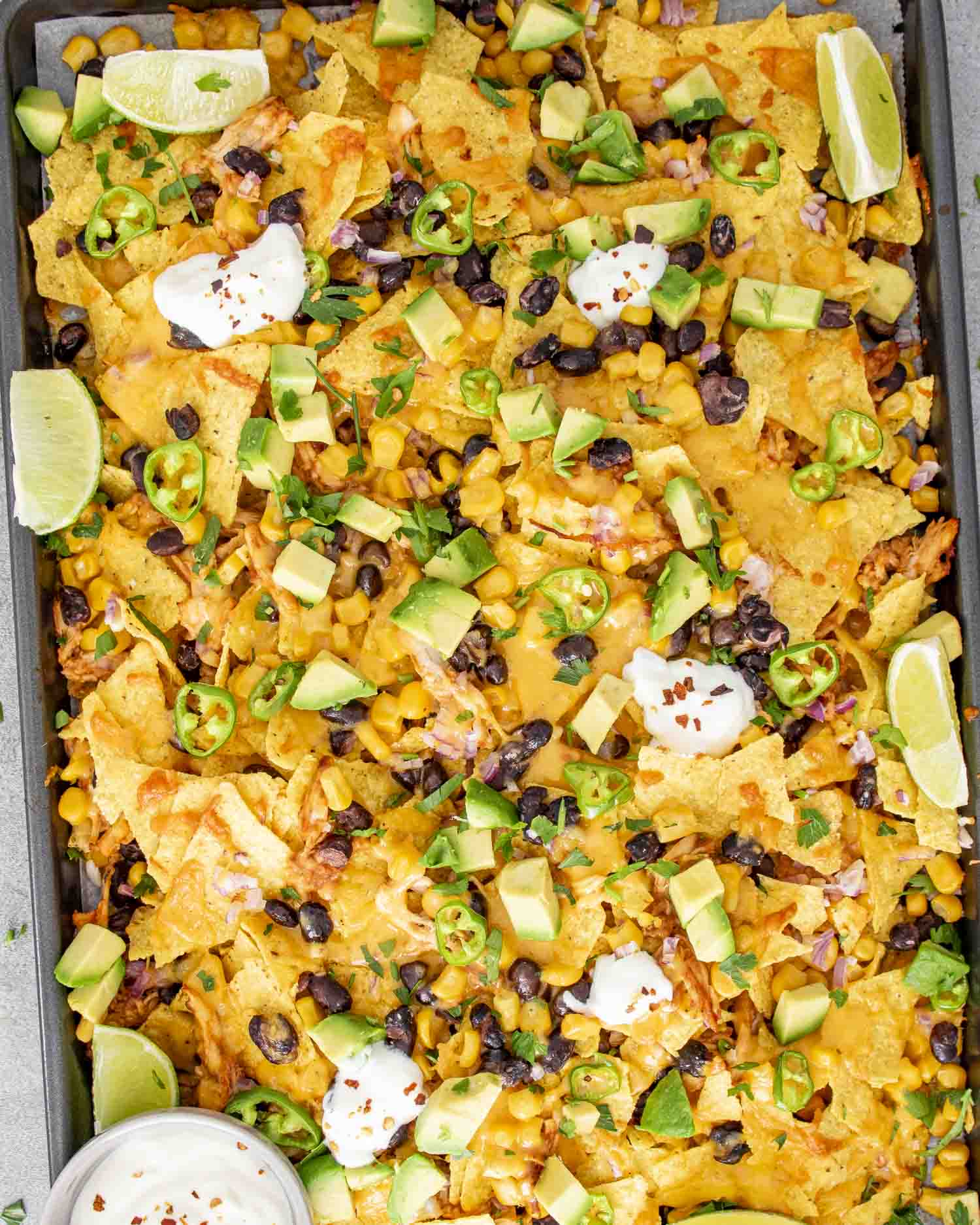 A sheet pan filled with BBQ chicken nachos topped with melted cheddar cheese, black beans, corn, diced avocado, jalapeño slices, fresh cilantro, and dollops of sour cream.