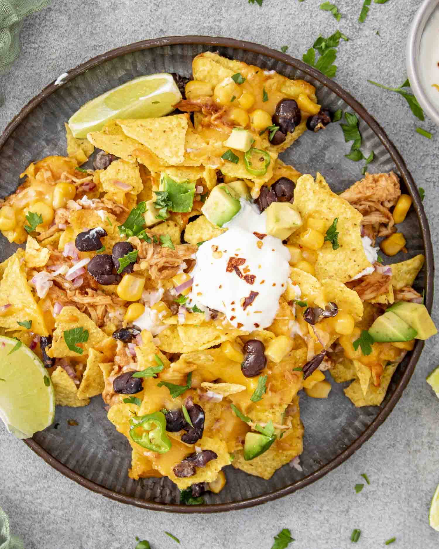 A plate of BBQ chicken nachos topped with melted cheese, black beans, corn, diced avocado, jalapeño slices, sour cream, and fresh cilantro. Lime wedges sit on the side for an extra burst of flavor.
