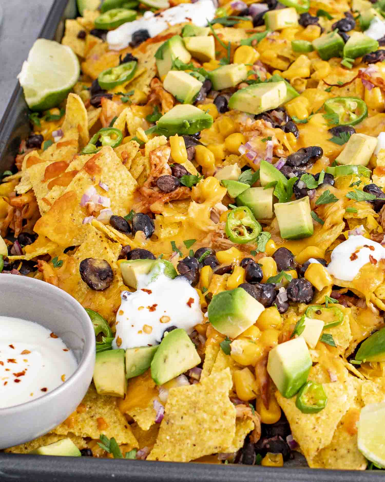 A sheet pan filled with BBQ chicken nachos topped with melted cheddar cheese, black beans, corn, diced avocado, jalapeño slices, fresh cilantro, and dollops of sour cream.