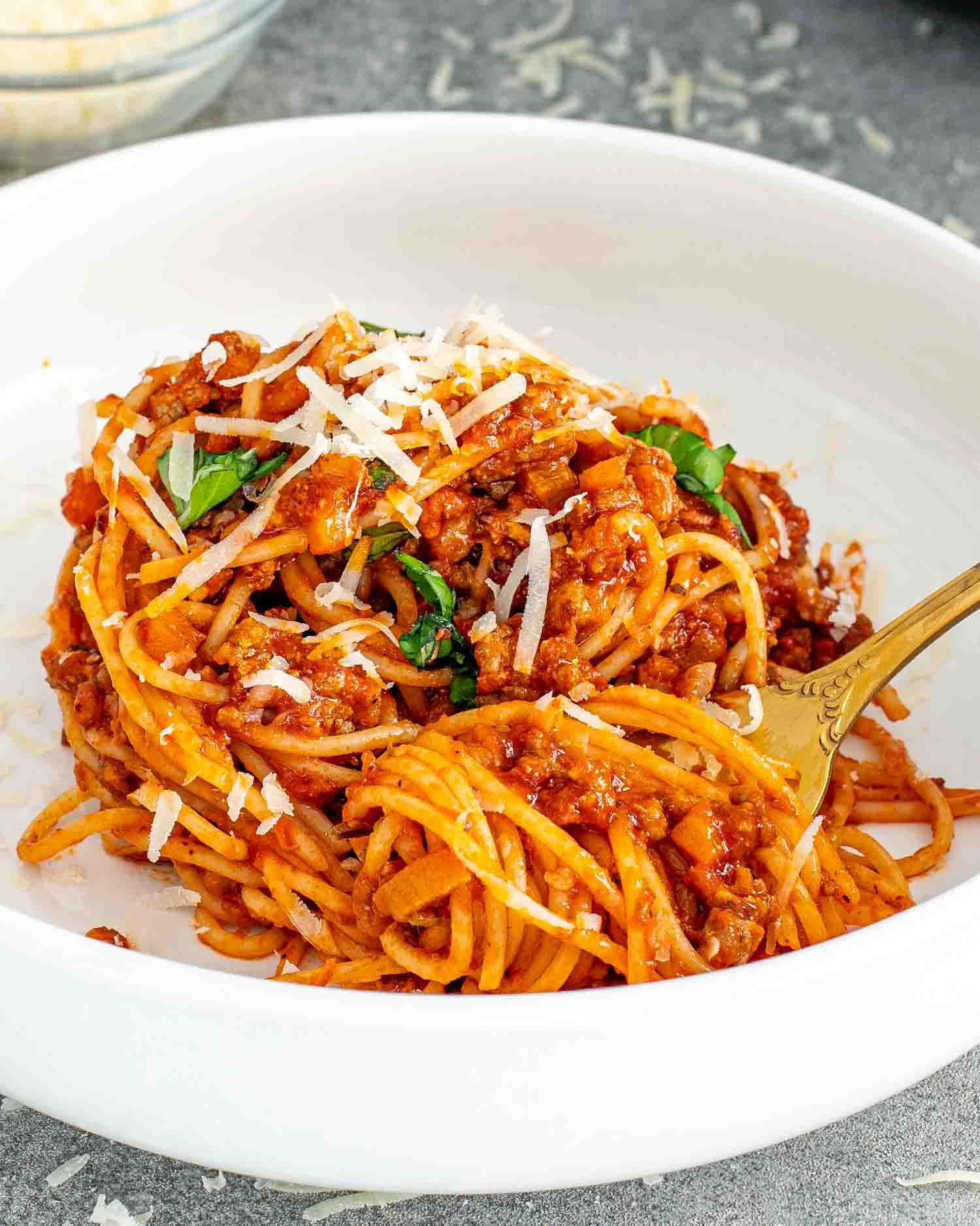 a white bowl with a serving of spaghetti bolognese in it, garnished with a bit of fresh basil and some grated parmesan cheese.