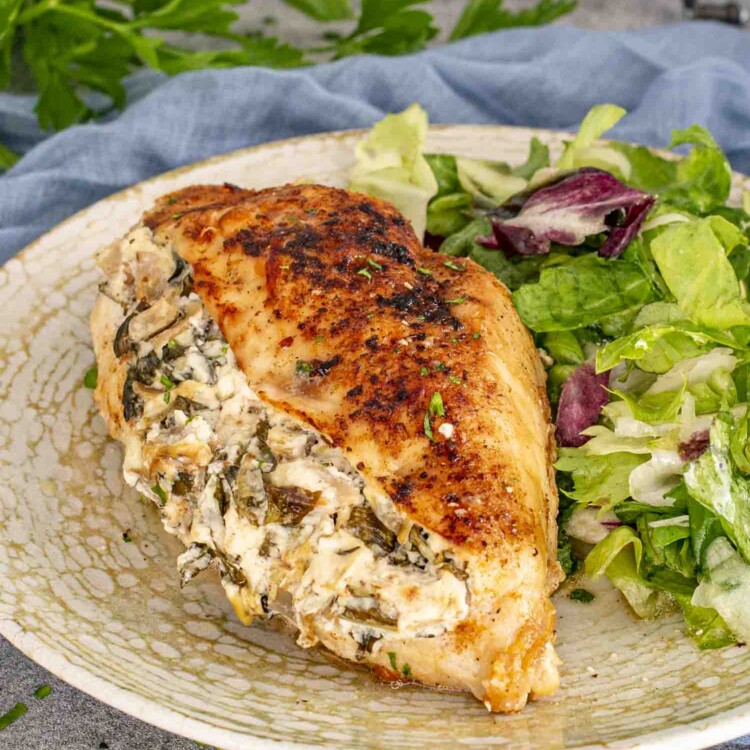 A stuffed chicken breast with a creamy spinach and artichoke filling, served with a fresh green salad on a speckled plate.
