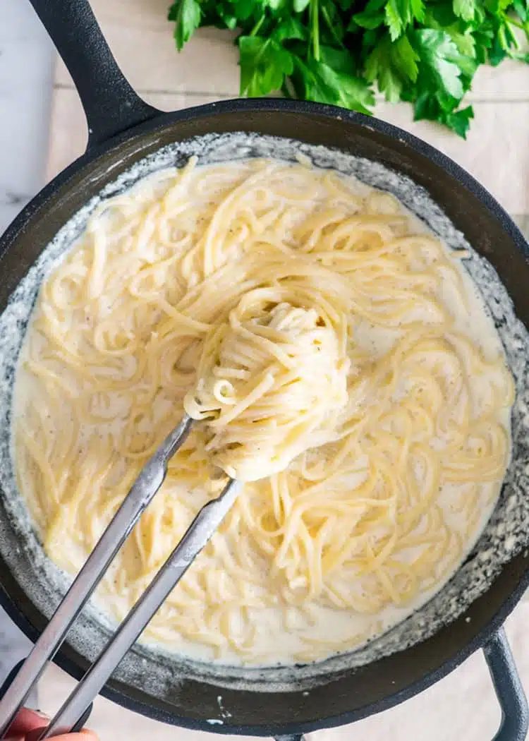 overhead shot of a pair of tongs lifting pasta alfredo out of a cast iron pan