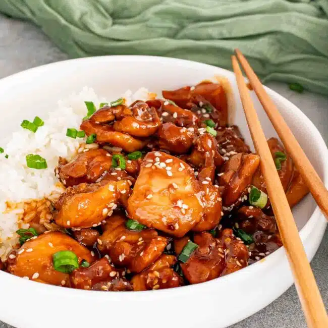 a serving of bourbon chicken with rice in a white plate garnished with sesame seeds and green onions.