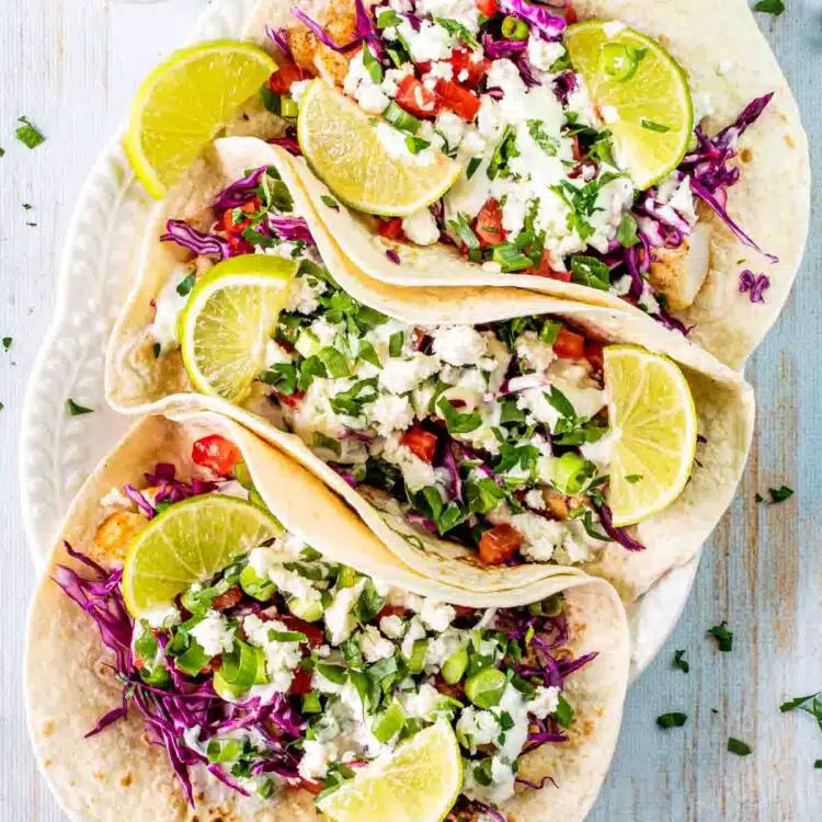 3 fish tacos with lime wedges on a serving platter.