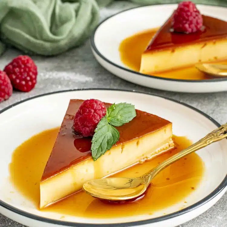 A silky smooth slice of flan topped with a fresh raspberry and mint leaf, resting in a pool of rich, golden caramel sauce on a white plate with a decorative gold spoon beside it. A simple, elegant dessert that looks as delicious as it tastes.