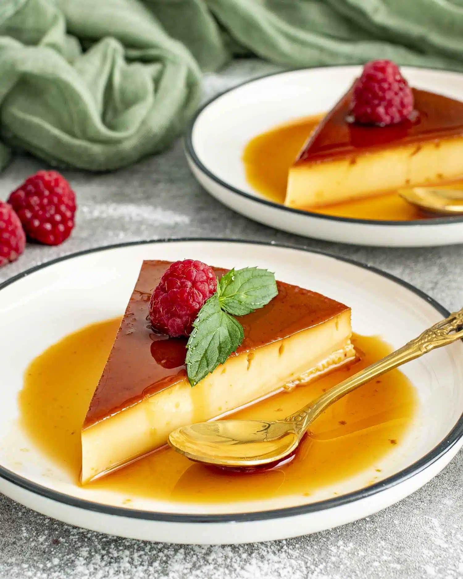 A silky smooth slice of flan topped with a fresh raspberry and mint leaf, resting in a pool of rich, golden caramel sauce on a white plate with a decorative gold spoon beside it. A simple, elegant dessert that looks as delicious as it tastes.