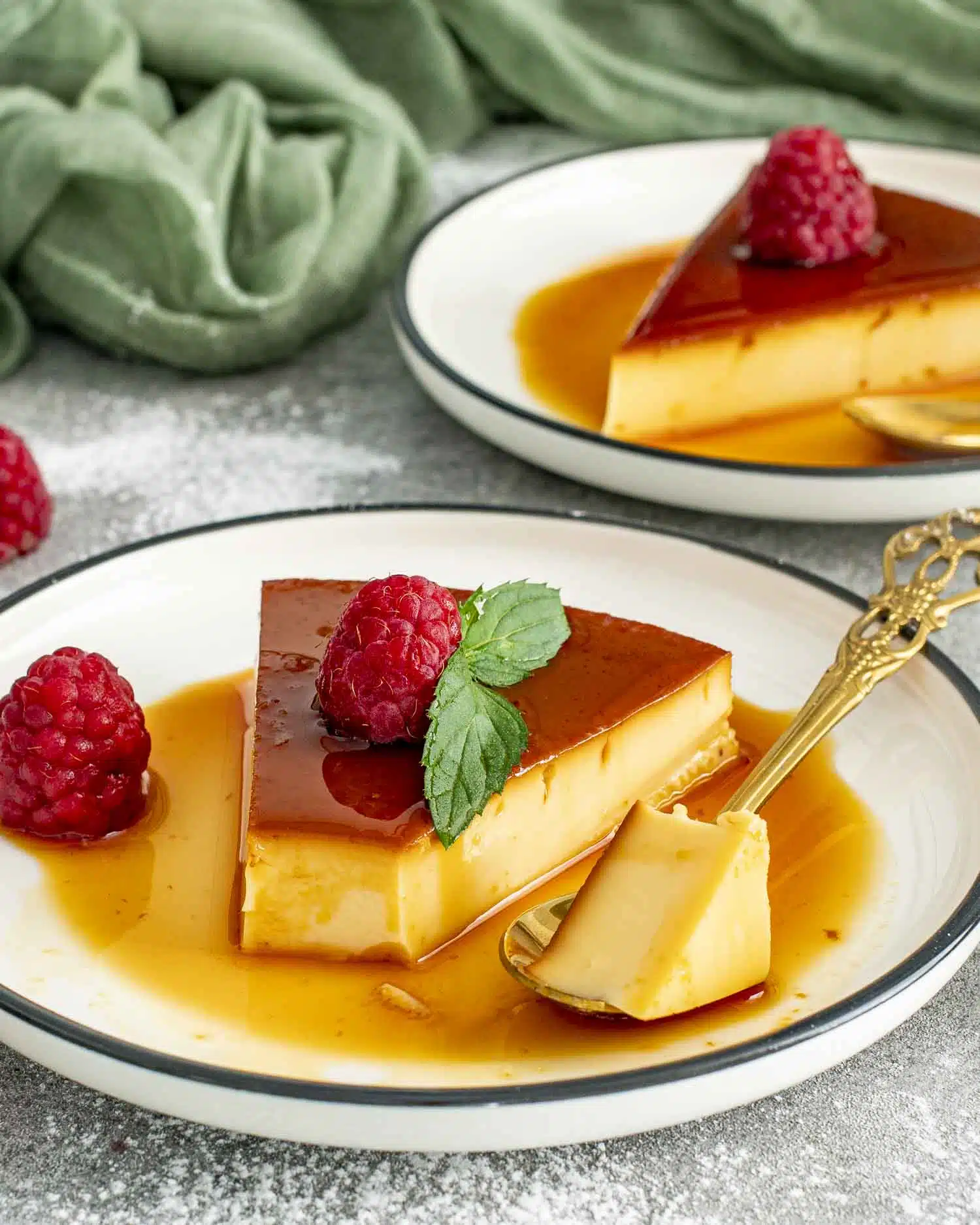 A silky smooth slice of flan topped with a fresh raspberry and mint leaf, resting in a pool of rich, golden caramel sauce on a white plate with a decorative gold spoon beside it.
