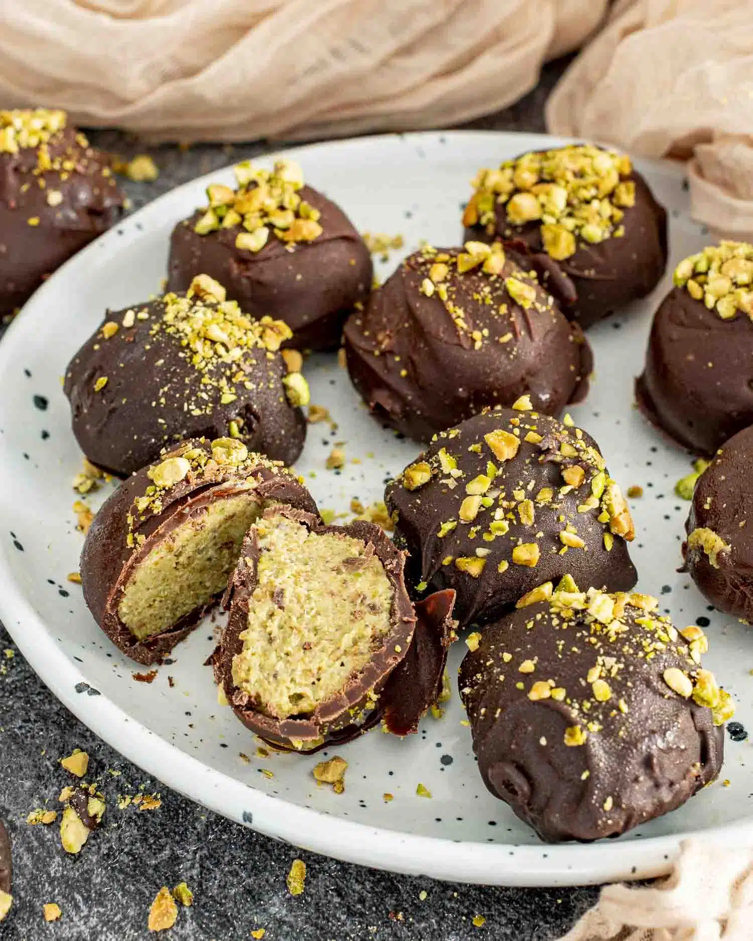 A white speckled plate filled with Frozen Pistachio and Date Chocolate Bites, coated in dark chocolate and sprinkled with crushed pistachios. One bite is cut open, revealing a creamy, nutty pistachio and date filling. Pistachio crumbs are scattered around the plate, set on a dark textured surface with soft beige fabric in the background.