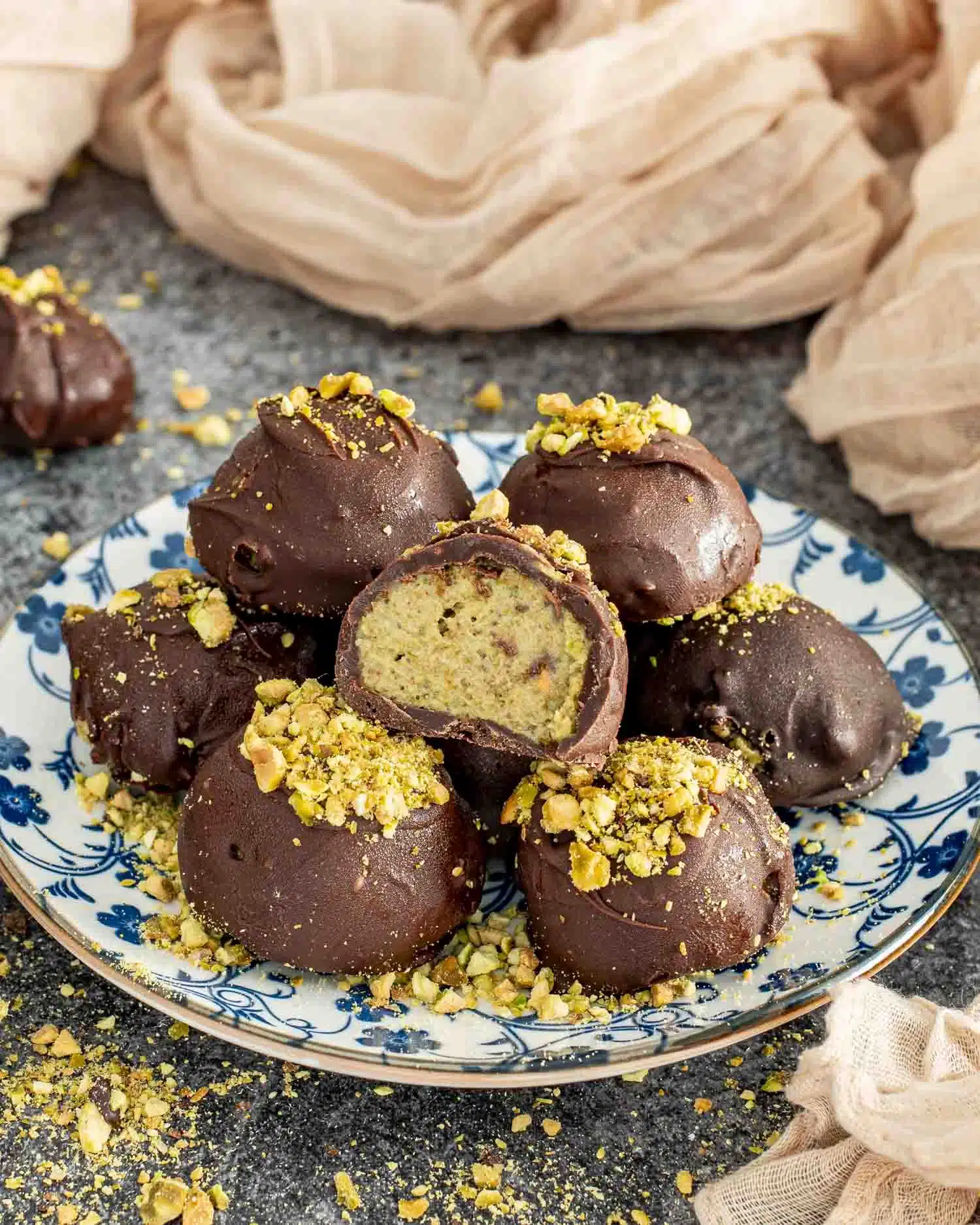 A plate of Frozen Pistachio and Date Chocolate Bites, coated in a glossy dark chocolate shell and topped with crushed pistachios. One bite is cut in half, revealing the creamy, nutty pistachio and date filling inside. The plate is set on a textured dark surface with scattered pistachio crumbs around it, with soft beige fabric in the background adding warmth to the scene.