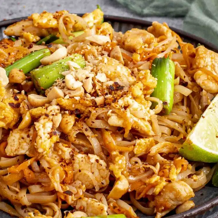 A plate of Pad Thai with rice noodles, tender chicken, scrambled eggs, green onions, and crushed peanuts. Chopsticks are lifting a portion of noodles, while a lime wedge adds a pop of color on the side.