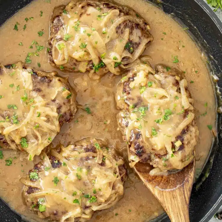 Juicy beef patties covered in rich, creamy onion gravy, garnished with fresh parsley, served hot in a black skillet.