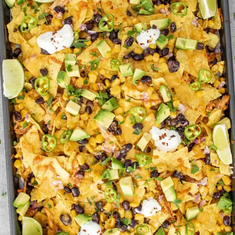 A sheet pan filled with BBQ chicken nachos topped with melted cheddar cheese, black beans, corn, diced avocado, jalapeño slices, fresh cilantro, and dollops of sour cream.